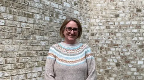 Emma Howgego/BBC Vicky Jenner stares at the camera. She wears black rimmed glasses, a beige jumper with a blue, white and gold pattern on the top half. She stands in front of a brick wall