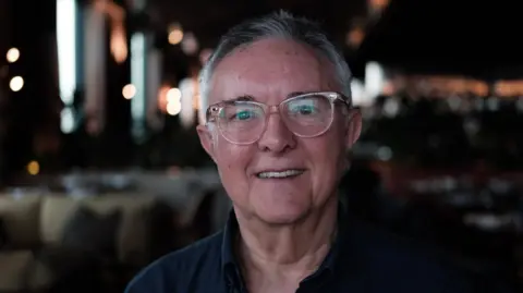 Ronnie Bergman looking directly at the camera and smiling. He has short grey hair and glasses, and is wearing a black shirt.