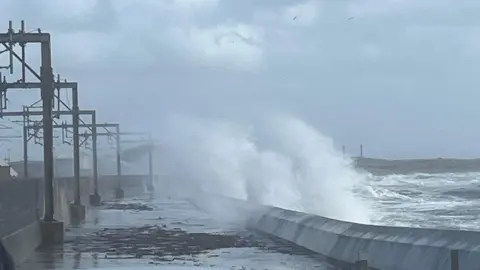 Waves can be seen crashing over the stone barrier onto a path next to a train track. In the background it is a cloudy day.