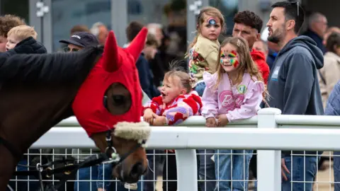 Bath Racecourse A horse with a red piece of fabric over the top of its head and ears is walking along a racecourse. A family watches on, with two girls with face paints on leaning on the racecourse's barrier.
