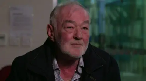 A man, with grey hair and a grey stubble, looking at the camera. He has a serious look on his face. He is wearing a shirt and a black jacket. The background of the image is blurred. 