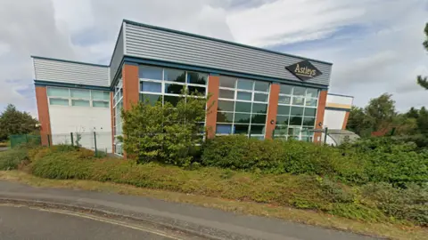 A distribution centre pictured from the road. There is a green fence and bushes in front of the building. A black diamond shape sign in the top right hand corner of the building reads "Astleys" in yellow writing. 