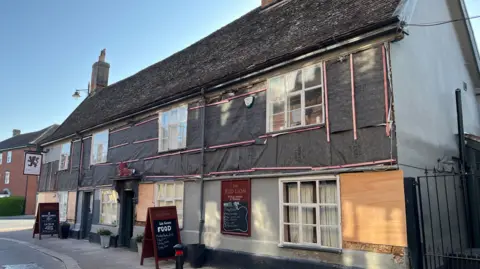 Darren Rozier/BBC A pub covered in tarpaulin on a street. A pub sign showing a Red Lion hangs from the left side of the building. Two boards advertising food are on the pavement outside the pub.