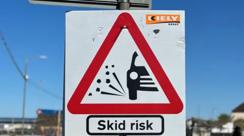 A road sign warning of a skid risk due to loose chippings. It features a red triangle with the black outline of a car with chip shooting upwards from the wheel. The words Sked risk are written in black below on a white background. Blue sky can be seen behind on a sunny day.