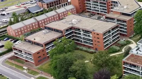BBC The image shows an aerial view of Bregawn House. Behind it balconies of Chatham House are visble and the corner of Carruthers Court. The buildings are made from similar materials with brick coloured walls and white balconies that span the width of the building. 