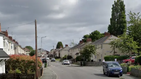 A google street view image of a residential street with houses on both side of the road. The sky is grey and cloudy