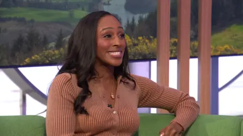 Alexandra Burke wears a brown long sleeved top and sits on the green One Show sofa