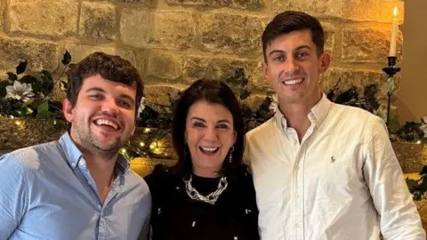 Harvey Isaac, Dawn Harper and Charlie Isaac are huddled together all looking at the camera with big smiles. They are pictured in front of a large Cotswold stone fireplace and chimney breast. The brothers both have dark short hair and are wearing button up shirts. Dawn is wearing a chunky silver necklace over a black embellished dress. The mantle piece is decorated with fairy lights, candles and festive foliage.