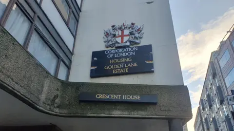 LDRS A close up of signage of Crescent House. Signs display the City of London's crest and the words “Corporation of London," "Housing," "Golden Lane Estate,” and below that "Crescent House".