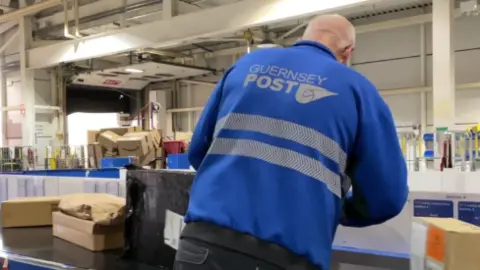 BBC A man wearing a Guernsey Post jacket putting parcels on the moving conveyor belt in the warehouse.