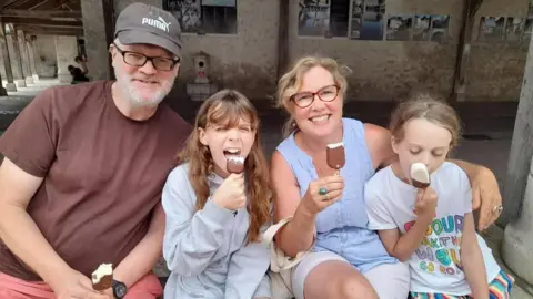 A man wearing a dark purple shirt, is sitting with a blonde woman wearing a blue shirt and white shorts. They are sitting with two blonde girls. They are all eating chocolate and vanilla ice cream.