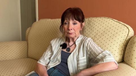 Yvonne Walton, a white woman in her 70s with dark red hair, she has a fringe and hair tied back, sitting on her yellow dotted sofa, wearing a pale green and white striped shirt jacket, grey top and blue jeans