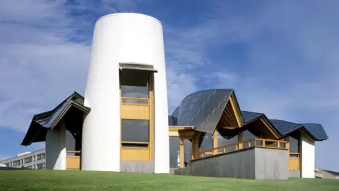 Maggie's Centre in Dundee. It has a bright white turret-style tower in the centre with a cottage-like building on either side with a wavy silver roof. It's a sunny day with blue sky and green grass in the foreground. 