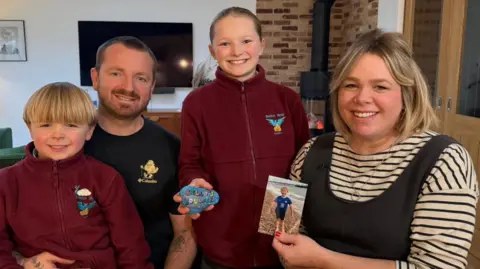 A man, woman, boy and girl smiling at the camera in a living room. The boy, on the far left, has short blonde hair and is wearing a burgundy fleece jacket. Behind him is a man with short brown hair who is wearing a black top. Next to him is a girl, who is standing up and wearing a burgundy fleece jacket. She is holding up a painted rock which has the words Billy's Boulders printed on it in rainbow letters. To her right is a woman who has chin-length blonde hair and is wearing a stripey long-sleeved t-shirt under dark-grey dungarees. She is holding up a photo of a boy wearing a blue football kit on a beach.