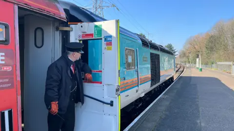 Tom Jackson/BBC A train driver wearing a black uniform and a cap, standing by the foot of the train door looking in the opposite direction of the camera. 