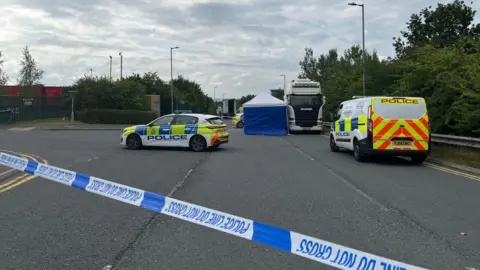 A police forensic tent stands next to a lorry at the side of the road. There are two police cars and a van alongside, with police tape in the foreground.