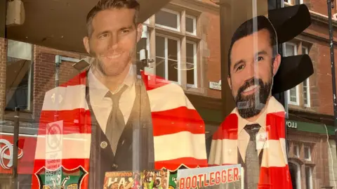 Cardboard cutouts of Ryan Reynolds and Rob McElhenney wearing Wrexham red and white striped scarves in a shop window.