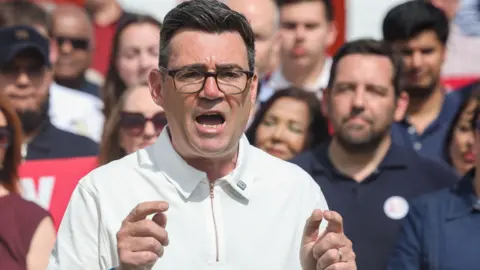 Andy Burnham, wearing a white T-shirt and glasses, speaks to a crowd