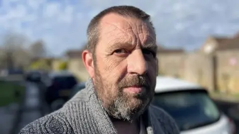 A close up of a man with short, brown hair and a beard. He is standing in front of a row of cars and is wearing a grey jumper.