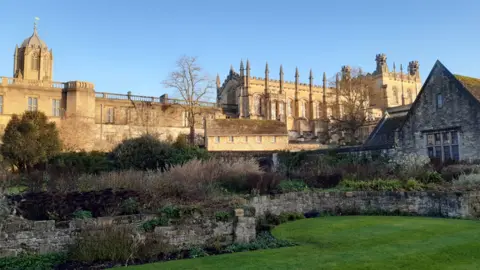 A cathedral stands in the background of the picture in a clear blue sky. In the foreground is a garden surrounded by a small stone wall with shrubbery behind it. There is another smaller stone building on the left of the picture. 