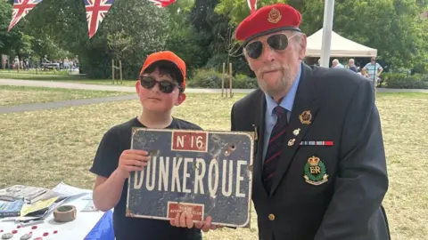Ben, standing next to a Royal British Legion man, who is wearing a jacket and red beret. Ben has on glasses and an orange cap and is holding a sign. They are in a field, with grass all around them. 
