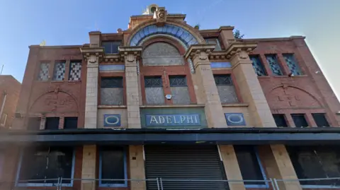 Google A large red brick building with stone accents, shutters covering doors and windows, and a security fence