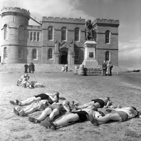 Getty Images Six people in shorts are lying on the grass in front of the castle. Further up towards the castle are two picnickers, while groups of people look at the Flora MacDonald statue