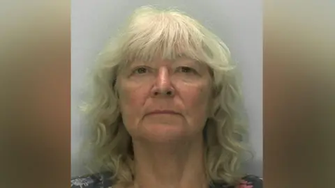 Gloucestershire Constabulary A mugshot of a woman in her 60s with wavy grey blonde hair and a fringe. She is straight faced as she stands in front of a pale grey background. From her shoulders, you can see she is wearing a floral T-shirt.