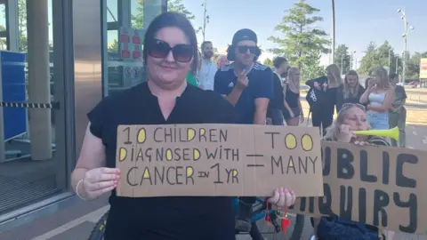 A woman is holding a cardboard banner saying too many children are being diagnosed with cancer. Another woman holds one calling for a public inquiry. Other people are gathered in the background.