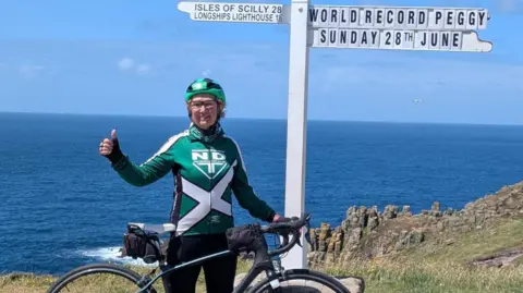 BBC A picture of Peggy Keenor-Crome at the finish line in Land's End. She is wearing a green cycling outfit, a green helmet and is posing with her bike.