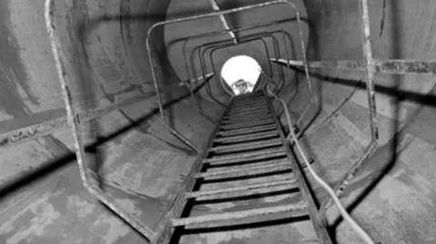 PA Media An old black and white picture from the bottom of a ladder in a drainage shaft, looking up towards the surface. A light-coloured wire is seen dangling down on the right. A person can be seen looking down from the top of the ladder.