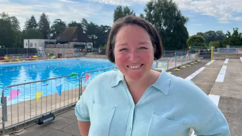 A woman standing beside an outdoor swimming pool, wearing a light blue collared top. The pool is lined with colourful bunting and safety barriers, with trees, buildings and a partly cloudy sky in the background.