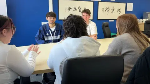 Five students sat around a board room table. You can see the backs of three female students and the front of two of the male students. They are all 17 years old.