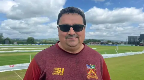 Norman Allen is wearing a red shirt with a red, green and blue logo on it. He has dark hair and is wearing black sunglasses. He is standing beside a cricket pitch.