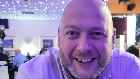 Kevin Rylance, 49, who has a bald head and a short beard, smiles at the camera 