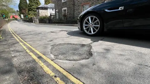 A pothole on a road in Tavistock which has been circled with white paint. There are double yellow lines and a navy blue car is driving past.