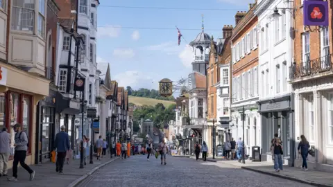 People walking on either side of a road in Guildford town centre. There is shops and businesses and blue skies. 