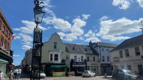 Edd Smith/BBC A general view of Fakenham town centre. Historic houses hold shops and cafes.
An ornate, black, Victorian-style lamp post is in the foreground. A few cars are parked on the road. People walk by. 