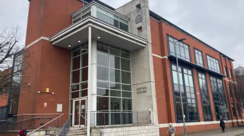 Warwick Crown Court, which is mainly brick-built, apart from the glass and concrete entrance is in the shot. A sign saying Justice Centre can be seen outside the doors. It is a cloudy day.
