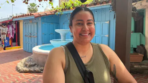 Rosa Cano, with dark hair pulled back and wearing an olive green tank top, stands smiling on a pedestrian street in front of a brightly painted, closed blue shop with stucco roof.