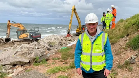 BBC Councillor John Murray standing on the shore at South Walney. He is wearing a white hard hat and a hi-vis jacket. Two diggers are moving rocks in the background.