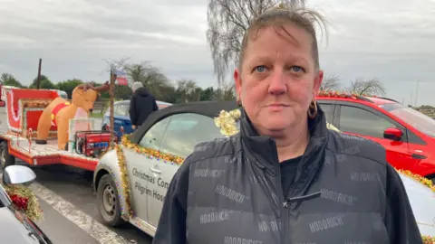 A woman looks at the camera with her hair tied back. She's wearing a black top, black body warmer and gold hoop earrings. Behind her are two stationary cars decorated with Christmas tinsel and a trialer carrying an inflatable reindeer.