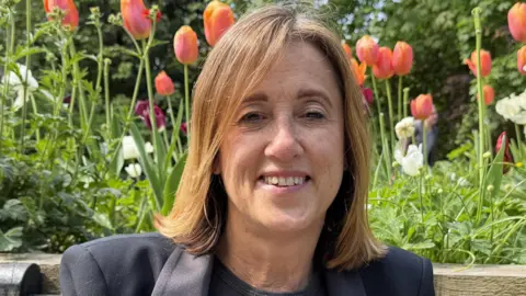 Jane Dodds looking into a camera. She has a dark jacket and black top, with flowers in the background.