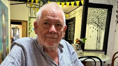 Chrissie Reidy/BBC Gatwick Area Conservation Campaign chairman Peter Barclay is wearing a striped shirt and sitting at a table with bunting above him and a picture on a wall next to him.