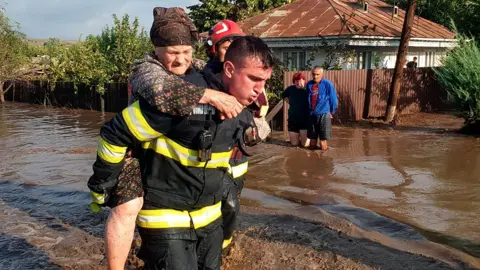 A Romanian rescuer carries an elderly woman on his back out of a flooded street, while a couple of neighbours look on in front of their house