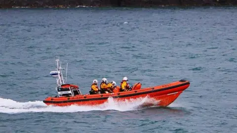 MARY DAVIES The new RNLI Lifeboat being delivered, it is a bright orange vessel, with people sat in bright yellow jackets and white helmets.