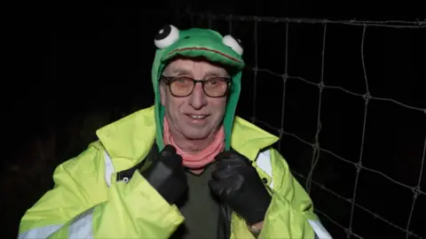 The image shows a person outdoors at night wearing a bright high‑visibility jacket and black gloves. The person is also wearing a green frog-themed hat with large cartoon eyes, standing next to a wire fence.