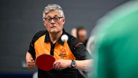Special Olympics a man in a Special Olympics Ulster jersey playing table tennis - the ball is visible but blurry because it is moving fast through the air, and his racket is poised
