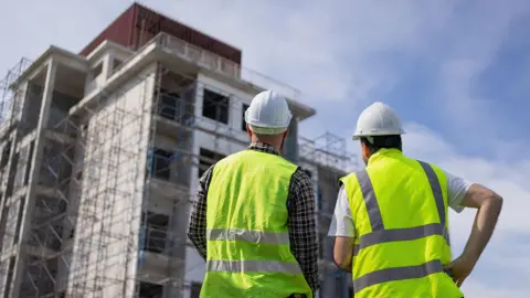 Getty Images Two men in hi-vis stand next to each other and look up to a  multi-storey residential building being constructed in front of them. The man on the left has a checked shirt, hi-vis and a white hard hat on. The man on the right has a white t-shirt on with his hi-vis and white hard hat.