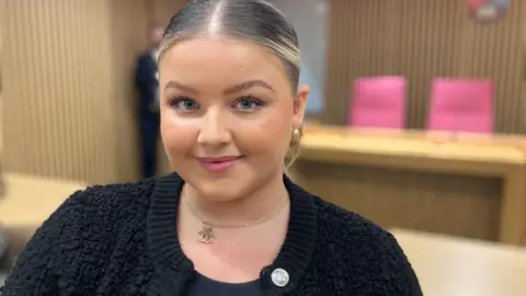 A blonde woman smiles at the camera, she has her hair tied back, is wearing make-up and pink lipstick, she has a gold chain around her neck and is wearing a black cardigan with large silver buttons.